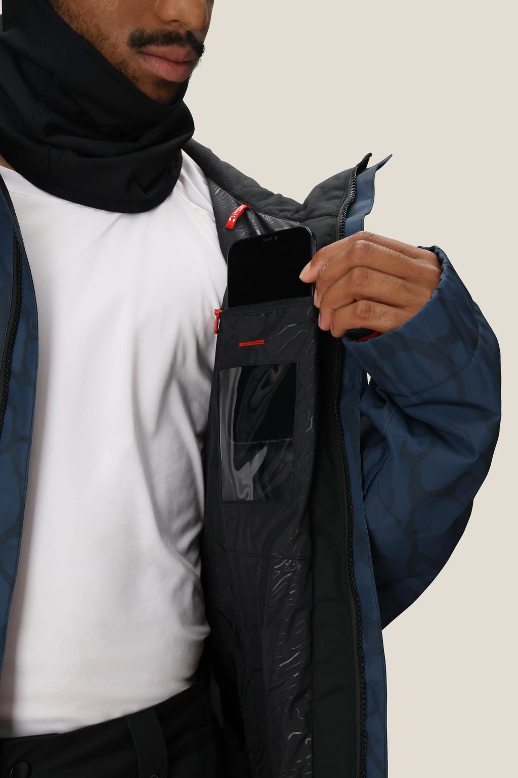 A person in a white shirt and blue jacket puts a smartphone into the inner chest pocket of the 686 Men's Foundation™ Insulated Jacket, partially unzipped to show its black lining and transparent ID slot.