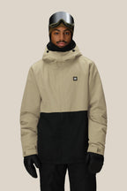 A person wearing the 686 Men's Foundation™ Insulated Jacket in tan and black, along with black gloves, pants, and a hood with ski goggles on their forehead, stands against a plain beige background.