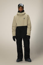 A person wearing black snow pants, black gloves, dark snow boots, ski goggles, and the 686 Men's Foundation™ Insulated Jacket in beige and black by 686 stands facing forward against a plain background.