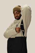 A person wearing a beige beanie and the 686 Men's Foundation™ Insulated Jacket in beige-and-black by 686 unzips a side zipper to show the jacket's inner lining, set against a plain light beige background.