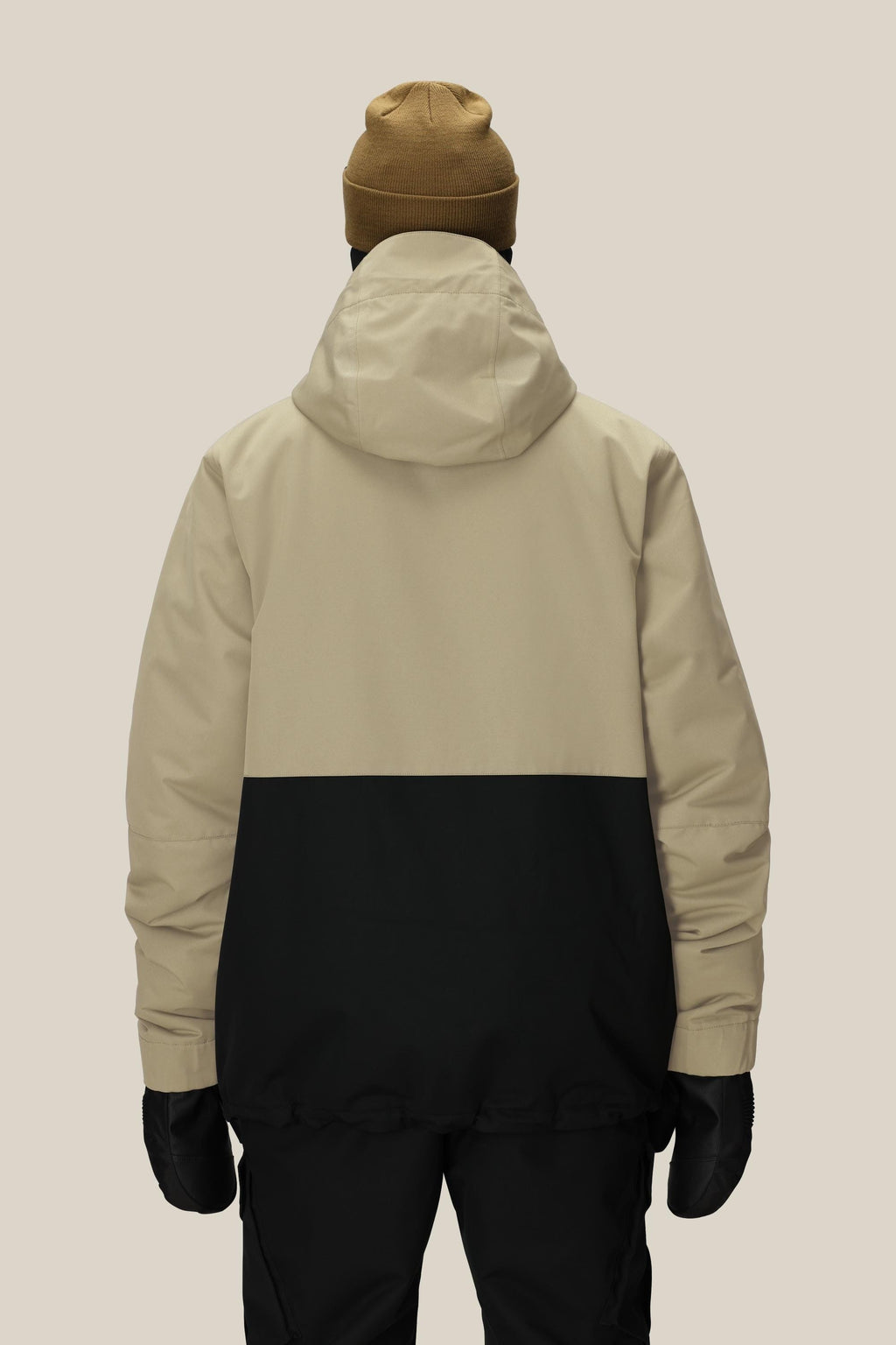 A person wearing a tan and black 686 Men's Foundation™ Insulated Jacket, black pants, black gloves, and a brown beanie stands facing away against a plain light background.