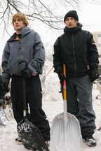 Two people in winter jackets and beanies stand outside in the snow holding large shovels. One wears the 686 Dojo® Jacket by 686 as they stand near a snow pile with bare tree branches overhead.