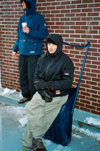 Two people are outside near a brick wall, dressed for winter. One, wearing the 686 Dojo® Jacket by 686, stands holding a cup while the other sits on a snow shovel. Snow and ice cover the ground around them.