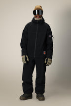A person stands facing forward wearing a black 686 Dojo® Jacket by 686, matching nylon waterproof pants, beige gloves, brown boots, a tan beanie, and large reflective ski goggles against a plain background.