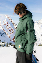 A man wearing the 686 Dojo® Jacket by 686 and black goggles stands on a snowy mountain, holding a colorful snowboard. Snow-covered peaks and a clear blue sky complete the stunning scene.