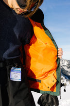 Wearing the 686 Dojo® Jacket with bright orange lining, a person shows its interior. A ski pass is clipped to their black snow pants, as they stand outdoors with snowy mountains and a blue sky behind them.
