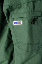 Close-up of the 686 Dojo® Jacket by 686, showing green nylon fabric with a subtle grid pattern, a visible pocket, black zipper pull, and a small rectangular patch featuring a blue and orange design.