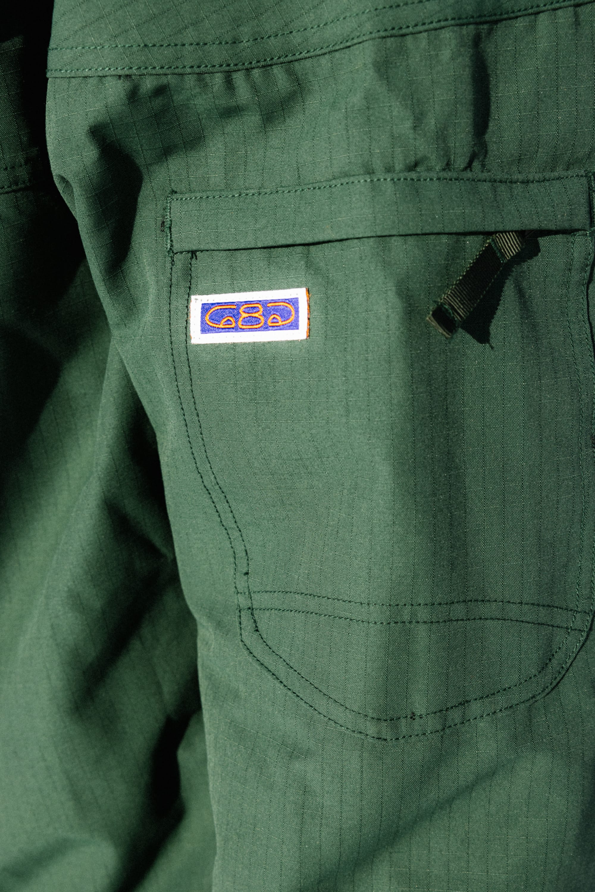 Close-up of the 686 Dojo® Jacket by 686, showing green nylon fabric with a subtle grid pattern, a visible pocket, black zipper pull, and a small rectangular patch featuring a blue and orange design.