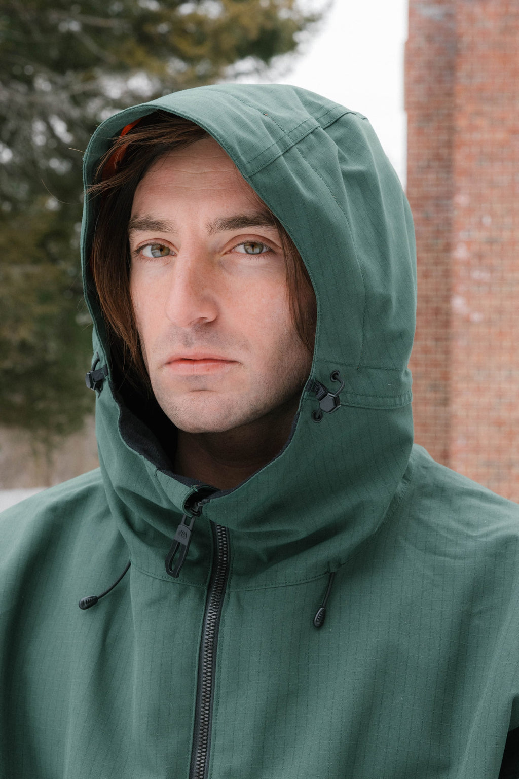 A person with long brown hair wears the 686 Dojo® Jacket by 686, featuring a green hood, while standing outdoors on a cloudy day. Trees and a brick wall appear in the background as they face the camera with a neutral expression.