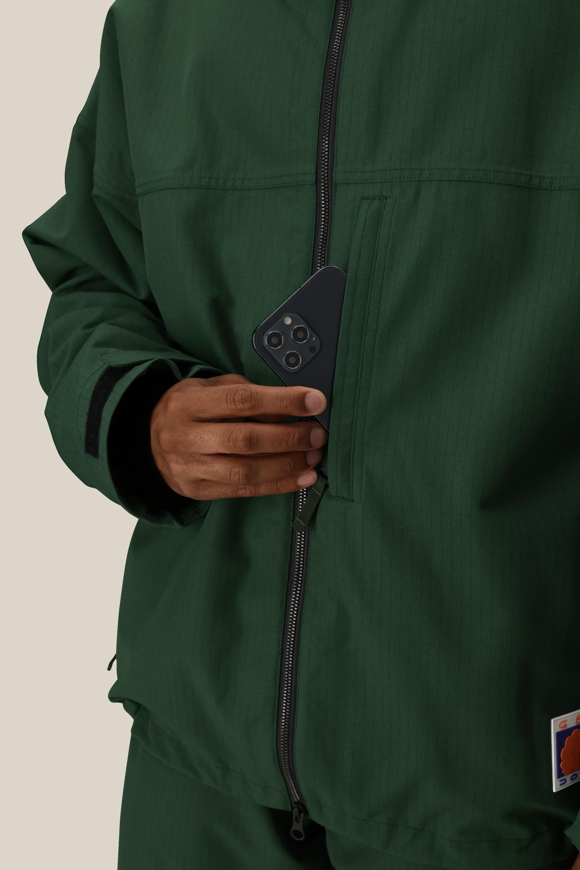 A person in a 686 Dojo® Jacket places a black smartphone into the zippered chest pocket. Only their hand and torso are visible, set against a neutral light background.