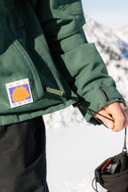 A person wearing the 686 Dojo® Jacket tightens the hem drawstring. A colorful patch decorates this men's nylon waterproof jacket by 686. Snowy mountains and bright sunlight appear in the background.