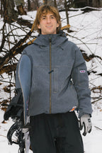 A person with light brown hair, dressed in the 686 Dojo® Jacket and black pants with gloves, stands outdoors in the snow holding a snowboard. Bare tree branches and snow are visible in the background.