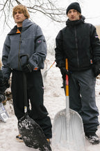Two young men stand in the snow, each with a shovel. They wear warm gloves, beanies, and the 686 Dojo® Jacket by 686. A mound of snow and bare tree branches behind them suggest a chilly winter day.