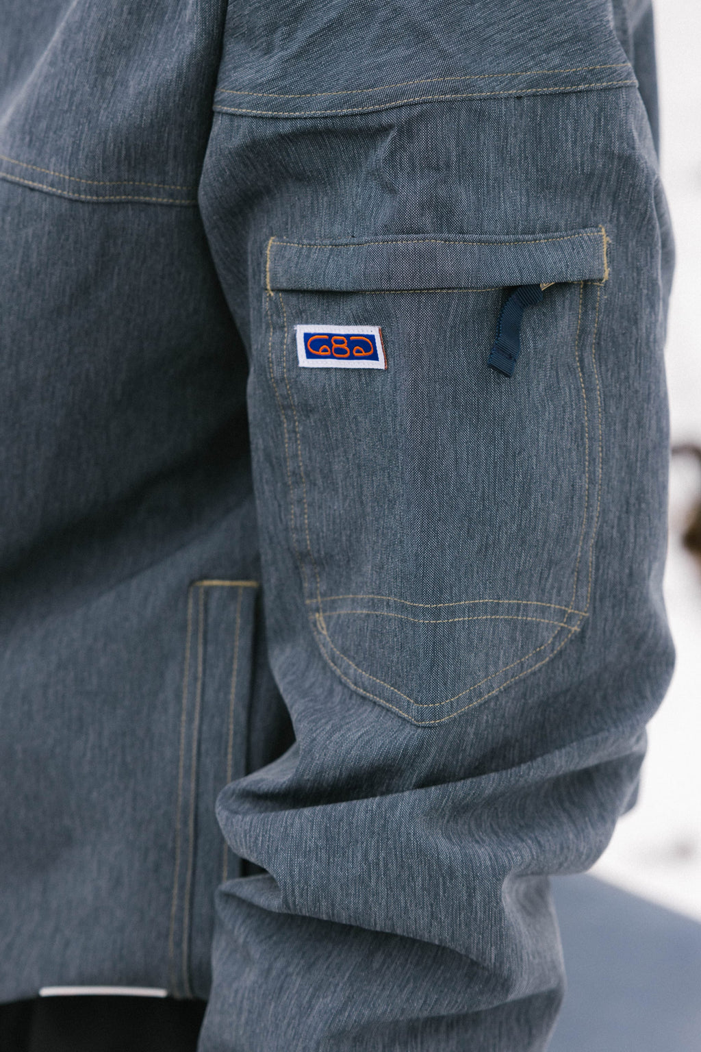 Close-up of the 686 Dojo® Jacket by 686, a men's blue-gray nylon jacket with yellow stitching, zippered sleeve pocket, and a small rectangular patch with a colorful logo above the pocket.