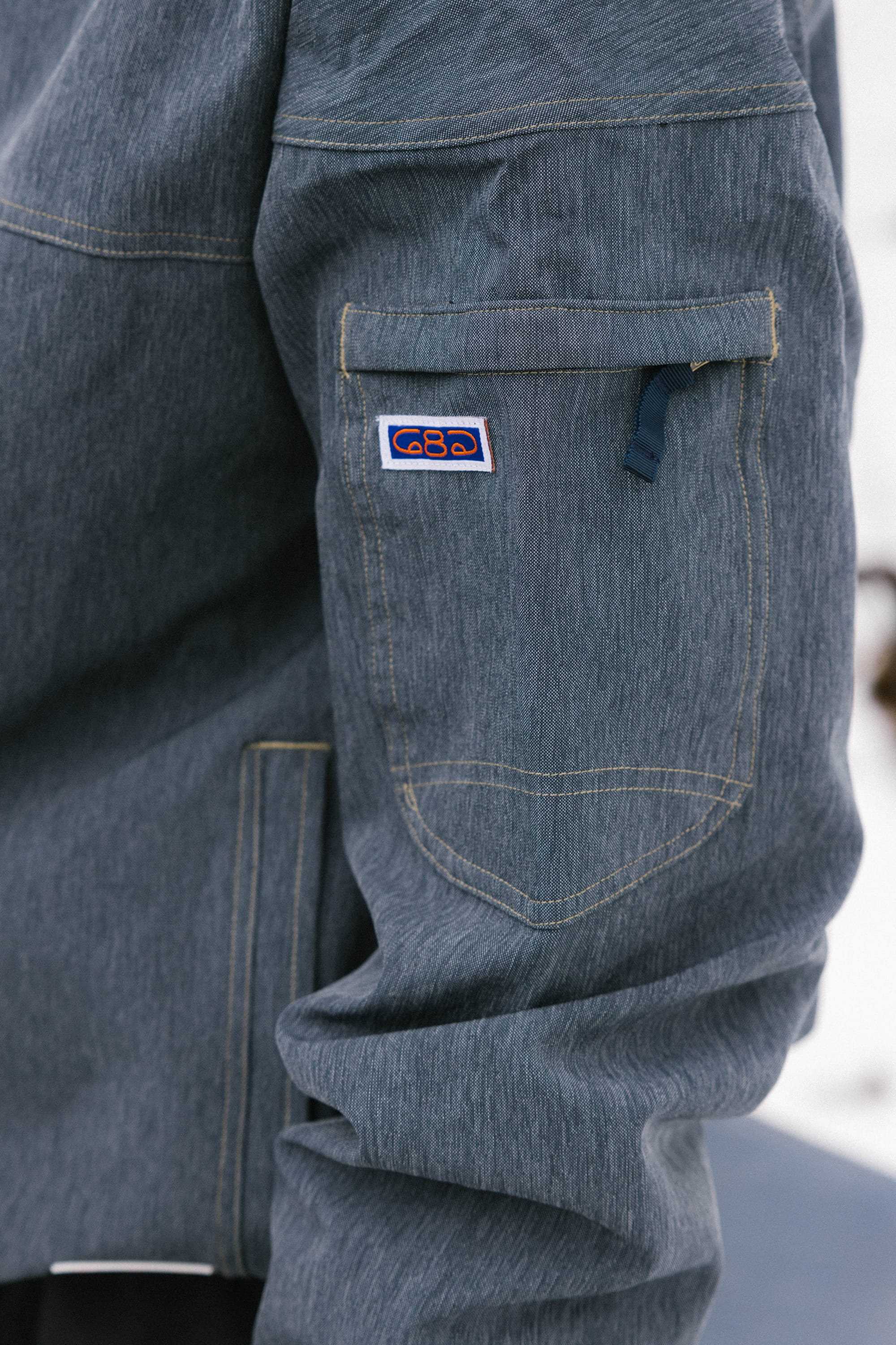 Close-up of the 686 Dojo® Jacket by 686, a men's blue-gray nylon jacket with yellow stitching, zippered sleeve pocket, and a small rectangular patch with a colorful logo above the pocket.