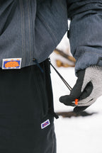 A person in the 686 Dojo® Jacket by 686 and patterned gloves tightens a drawstring on their black snow pants, with snow visible on the ground in the background.