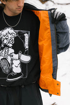 A person wearing a 686 Dojo® Jacket by 686 with an orange lining, black gloves, a chain necklace, and a black t-shirt featuring a cartoon angry character holding a bat, with snow visible in the background.