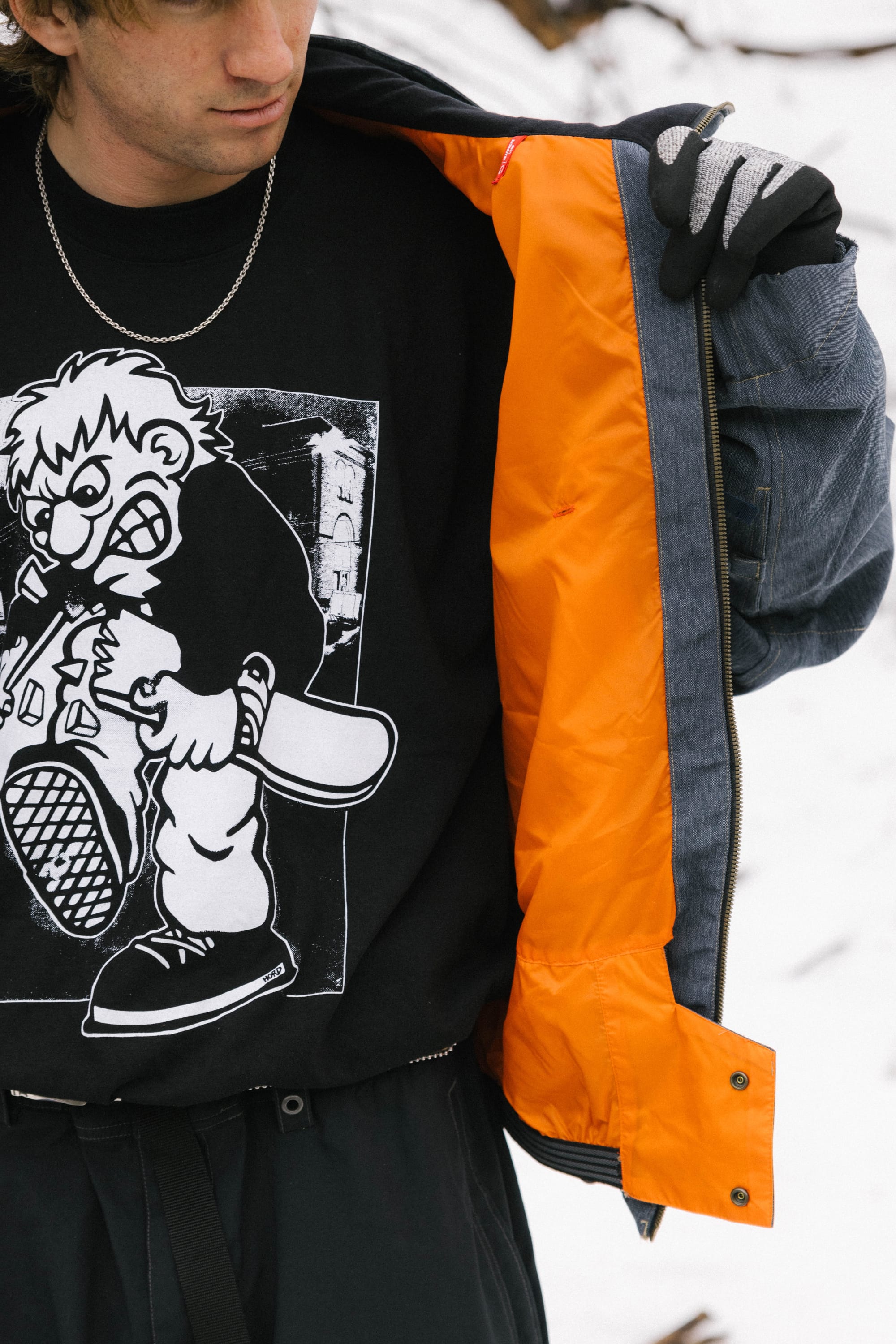 A person wearing a 686 Dojo® Jacket by 686 with an orange lining, black gloves, a chain necklace, and a black t-shirt featuring a cartoon angry character holding a bat, with snow visible in the background.