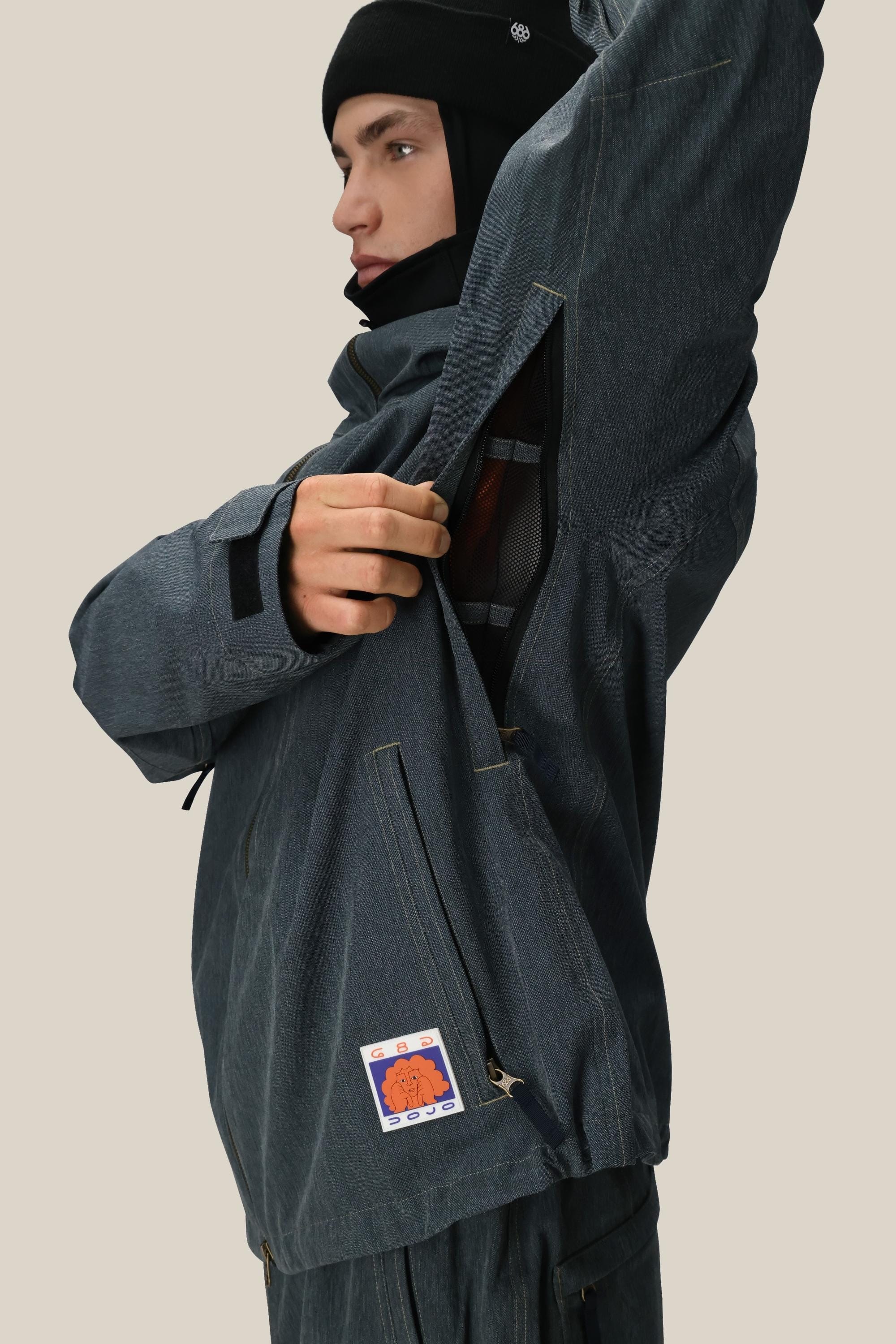 Someone in the 686 Dojo® Jacket by 686, colored dark gray, unzips a large side vent to show its mesh lining. A colorful patch is visible near the lower zipper. The person also wears a black beanie against a light beige background.