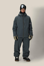 A person faces forward in a gray 686 Dojo® Jacket by 686, matching loose pants, gloves, black ski goggles, a black hat, and black sneakers with white soles against a plain beige background.