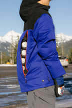 A person wears the 686 Mens PBR Cooler Insulated Jacket with a side zipper open, showing several cans stored inside. Snowy mountains and trees appear in the background.