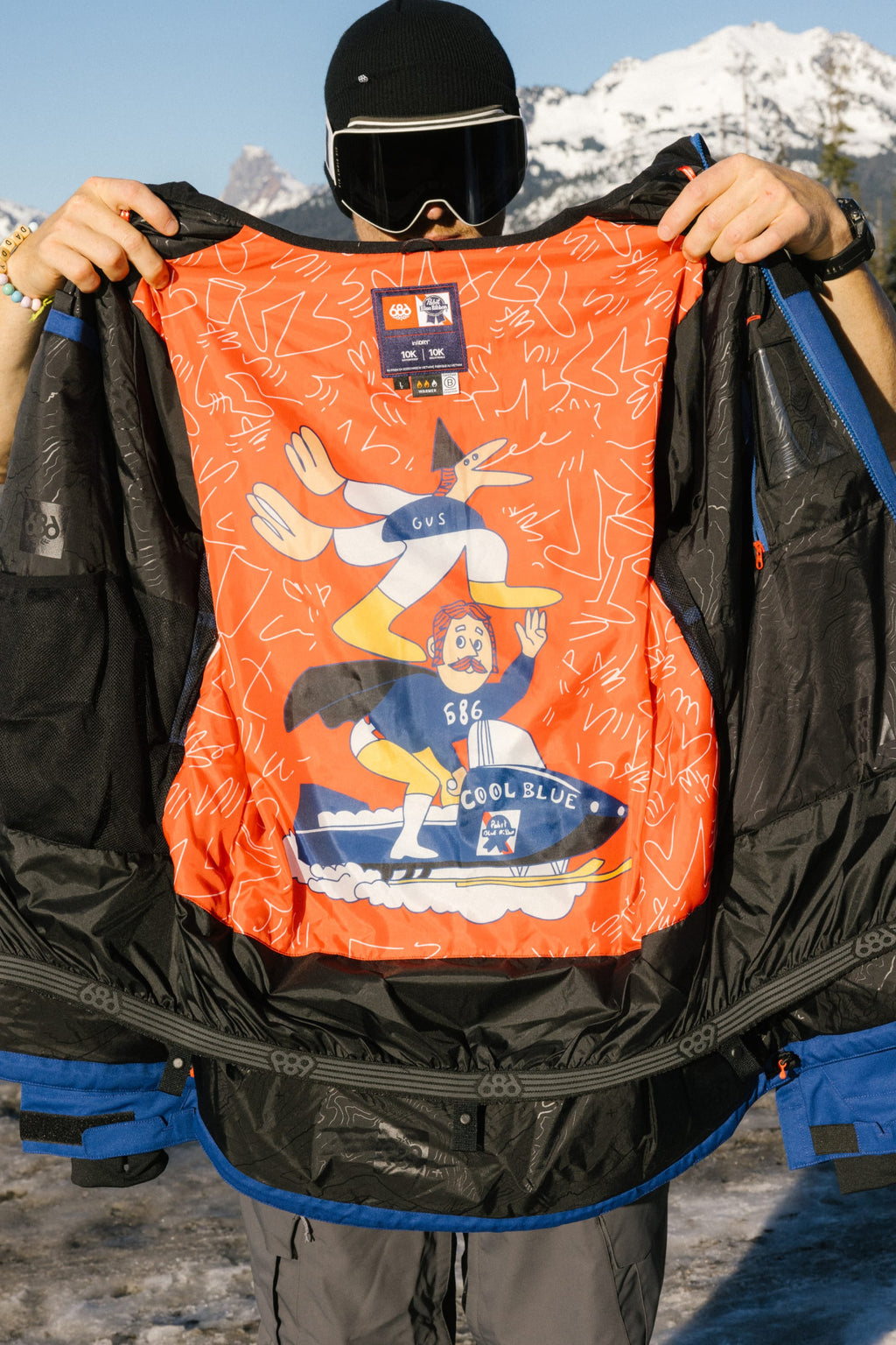 A person in winter gear shows off the bright red lining of the 686 Mens PBR Cooler Insulated Jacket, featuring playful graphics and text. Snowy mountains provide a scenic backdrop, highlighting the jackets style and warmth by 686.