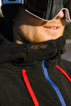 Close-up of a person wearing the 686 Mens PBR Cooler Insulated Jacket with red and blue drawstrings, black ski goggles, and a beanie. Their light reddish-brown beard and embroidered collar text are visible. Brand: 686.