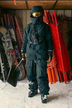 Wearing a 686 Men's GORE-TEX GT Thermagraph® Jacket, a person stands indoors holding a snow shovel. Snowboards, red poles, orange safety nets, and snow are visible in the background.