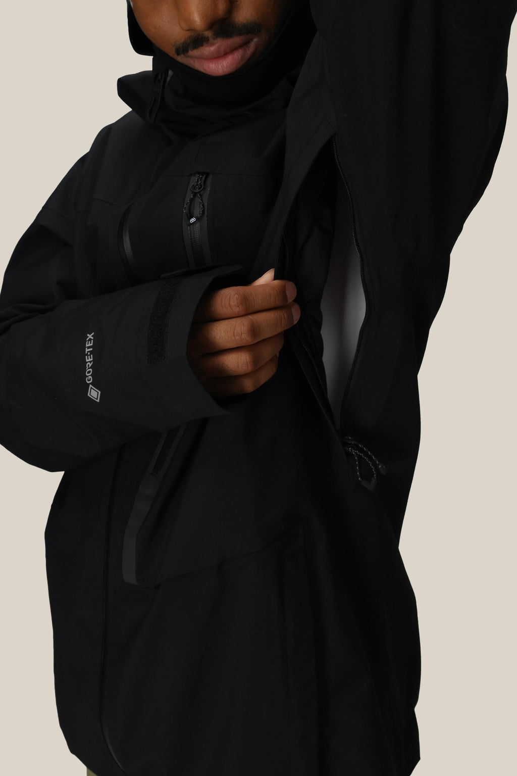 A person in the 686 Men's GORE-TEX GT Thermagraph® Jacket by 686 unzips the underarm vent, showing a white inner layer. Their face is partly visible, with a plain beige background.
