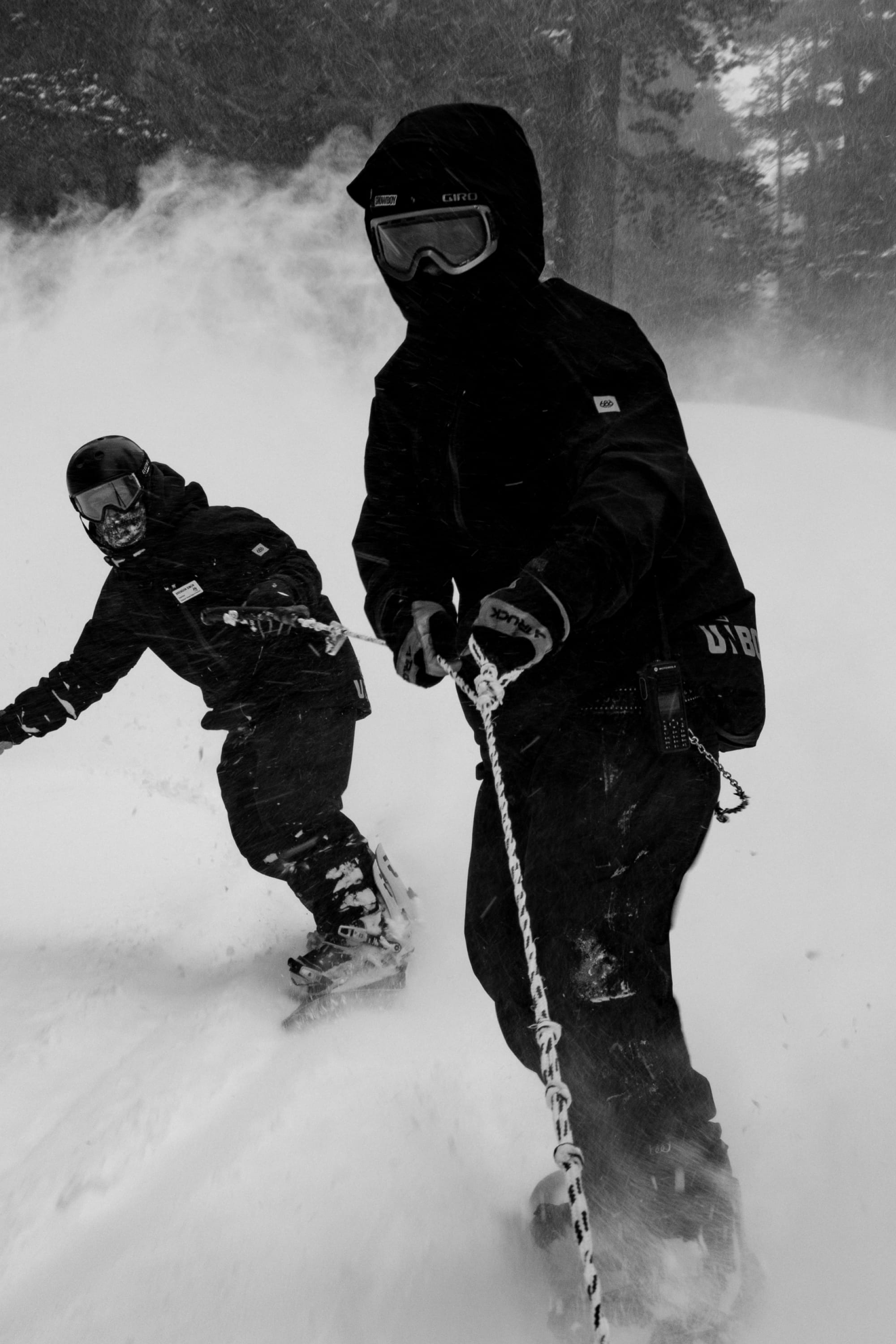 Two people in 686 Men's GORE-TEX GT Thermagraph® Jackets snowboard through deep snow, holding a rope as snow swirls around them in a black-and-white, forested scene.