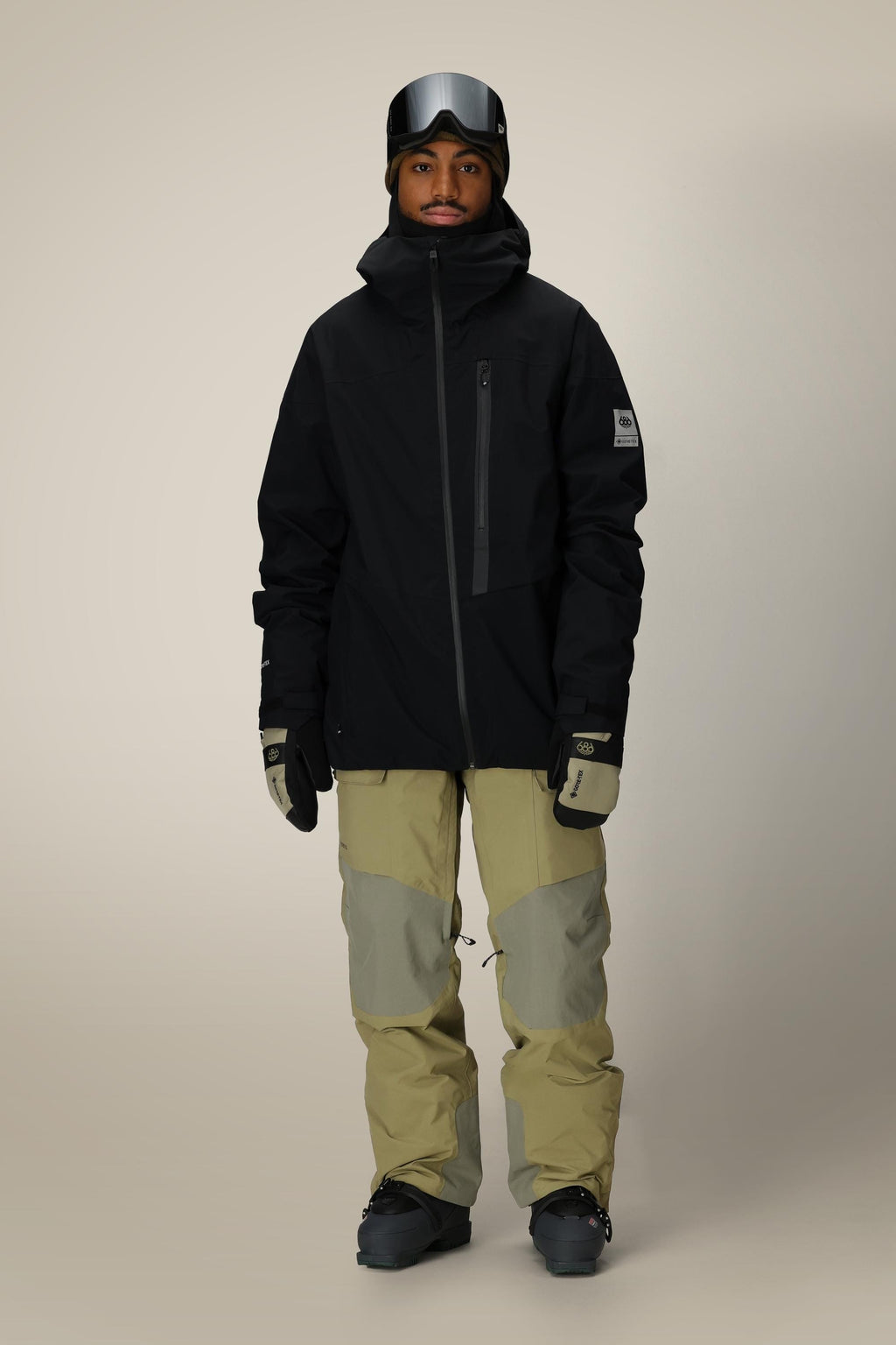A person wearing the 686 Men's GORE-TEX GT Thermagraph® Jacket in black stands facing forward, paired with light tan and gray snow pants, gloves, black ski boots, and ski goggles on their head against a plain beige background.