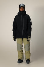 A person wearing the 686 Men's GORE-TEX GT Thermagraph® Jacket in black stands facing forward, paired with light tan and gray snow pants, gloves, black ski boots, and ski goggles on their head against a plain beige background.
