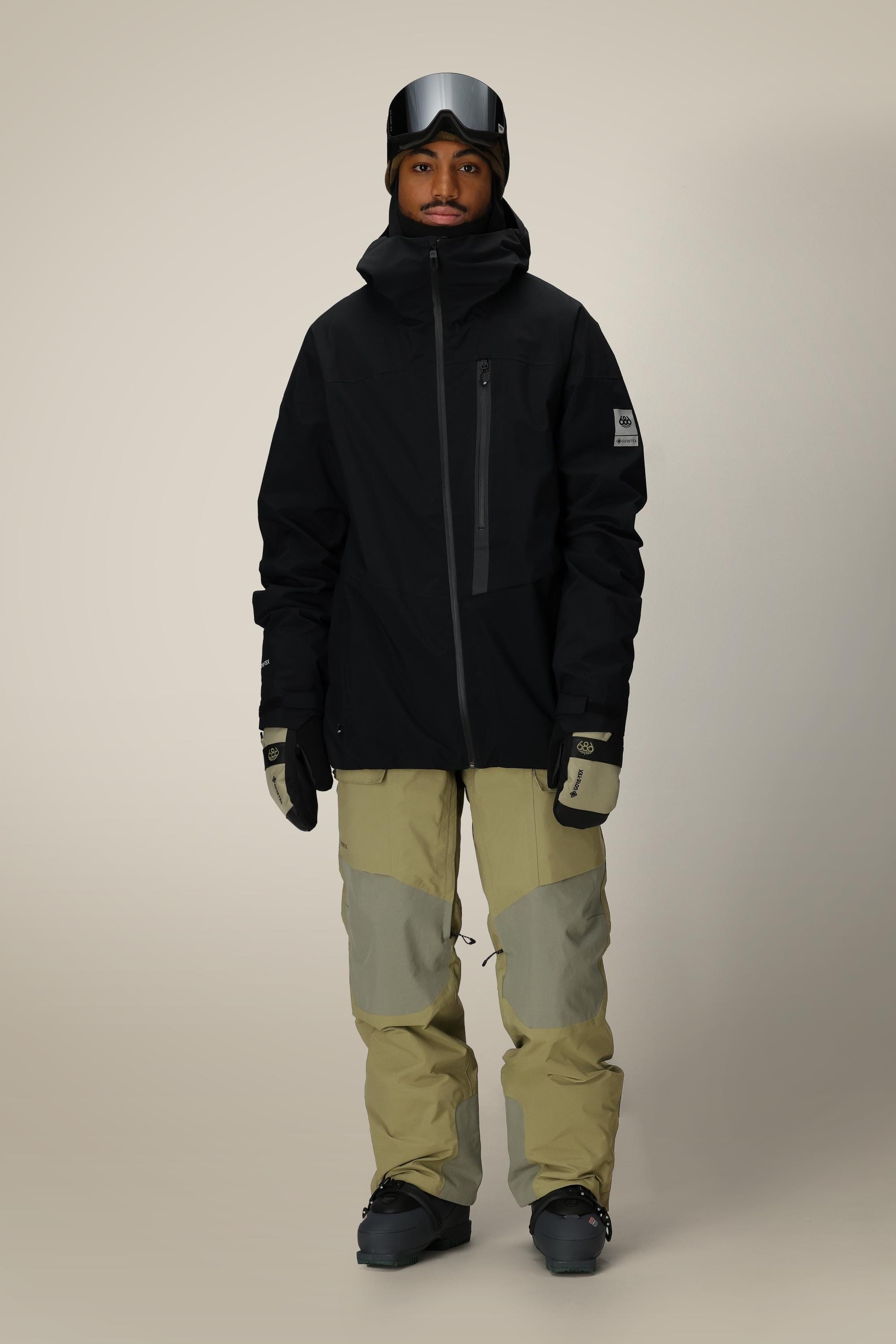 A person wearing the 686 Men's GORE-TEX GT Thermagraph® Jacket in black stands facing forward, paired with light tan and gray snow pants, gloves, black ski boots, and ski goggles on their head against a plain beige background.