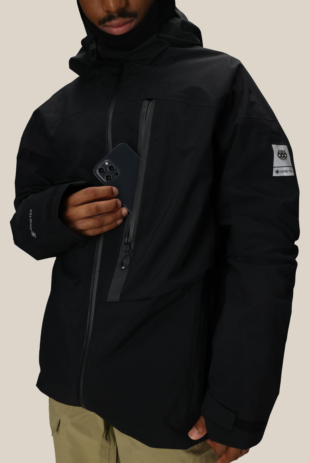 A person in a 686 Men's GORE-TEX GT Thermagraph® Jacket holds a smartphone partially tucked into the chest pocket. The black waterproof jacket features a patch on the upper arm, tan pants, and multiple zippered pockets.