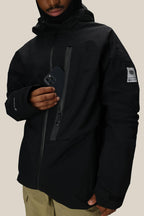 A person in a 686 Men's GORE-TEX GT Thermagraph® Jacket holds a smartphone partially tucked into the chest pocket. The black waterproof jacket features a patch on the upper arm, tan pants, and multiple zippered pockets.