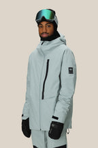 A person wears the 686 Men's GORE-TEX GT Thermagraph® Jacket, black gloves, a black beanie, and reflective ski goggles while standing against a plain beige background.