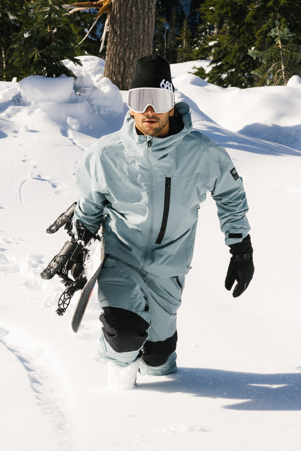 Wearing the 686 Men's GORE-TEX GT Thermagraph® Jacket, a person strides through deep snow among snow-covered trees, carrying a snowboard under one arm and wearing black gloves and white goggles.