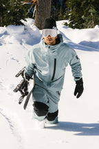 Wearing the 686 Men's GORE-TEX GT Thermagraph® Jacket, a person strides through deep snow among snow-covered trees, carrying a snowboard under one arm and wearing black gloves and white goggles.