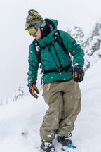 Wearing the 686 Men's GORE-TEX GT Thermagraph® Jacket, a person stands on a snowy slope among snow-covered trees, dressed in winter gear with tan pants, gloves, goggles, and a beanie.