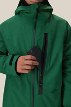A person in a green 686 Men's GORE-TEX GT Thermagraph® Jacket with a hood partially unzips the waterproof chest pocket and puts a black smartphone inside. The 686 jacket features Velcro cuffs and is set against a plain, light background.
