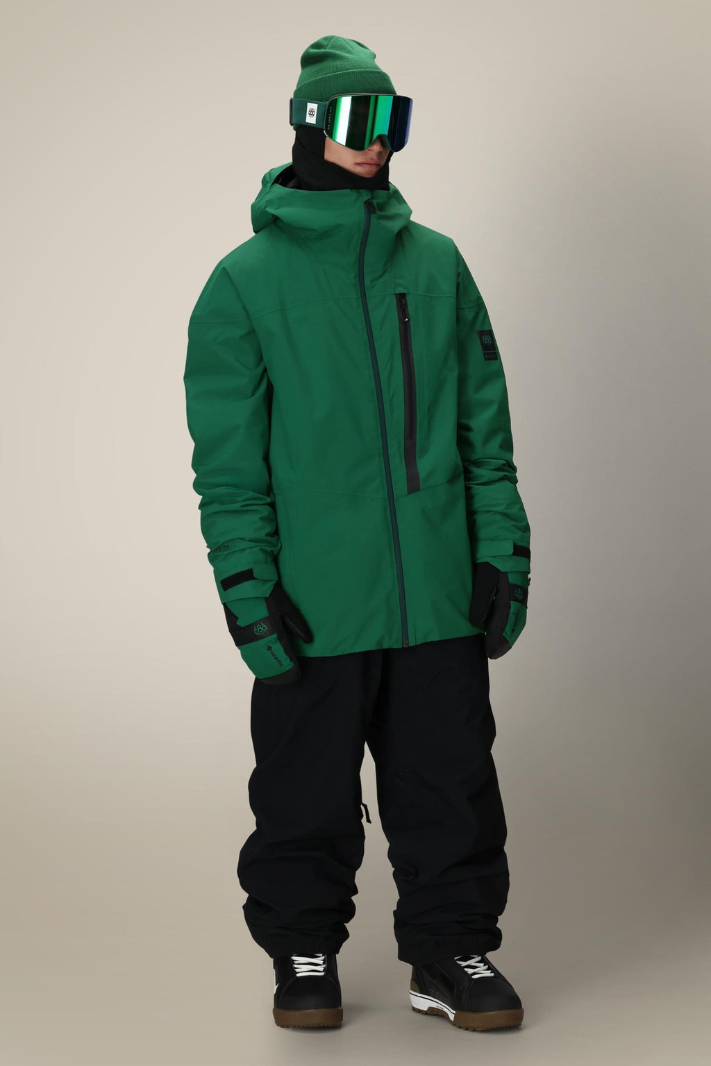 A person wearing the 686 Men's GORE-TEX GT Thermagraph® Jacket, green gloves, beanie, and ski goggles—paired with black snow pants and boots—stands facing slightly left against a neutral background.