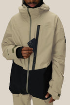 A person wearing the 686 Men's GORE-TEX GT Thermagraph® Jacket in beige and black places a black smartphone into its vertical zippered chest pocket. The 686 jacket features waterproof nylon, a high collar, and partially reveals the wearer’s face.