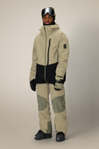 A person stands facing forward against a plain background, wearing the 686 Men's GORE-TEX GT Thermagraph® Jacket in beige and black, paired with matching snow pants, gloves, black snow boots, and ski goggles.