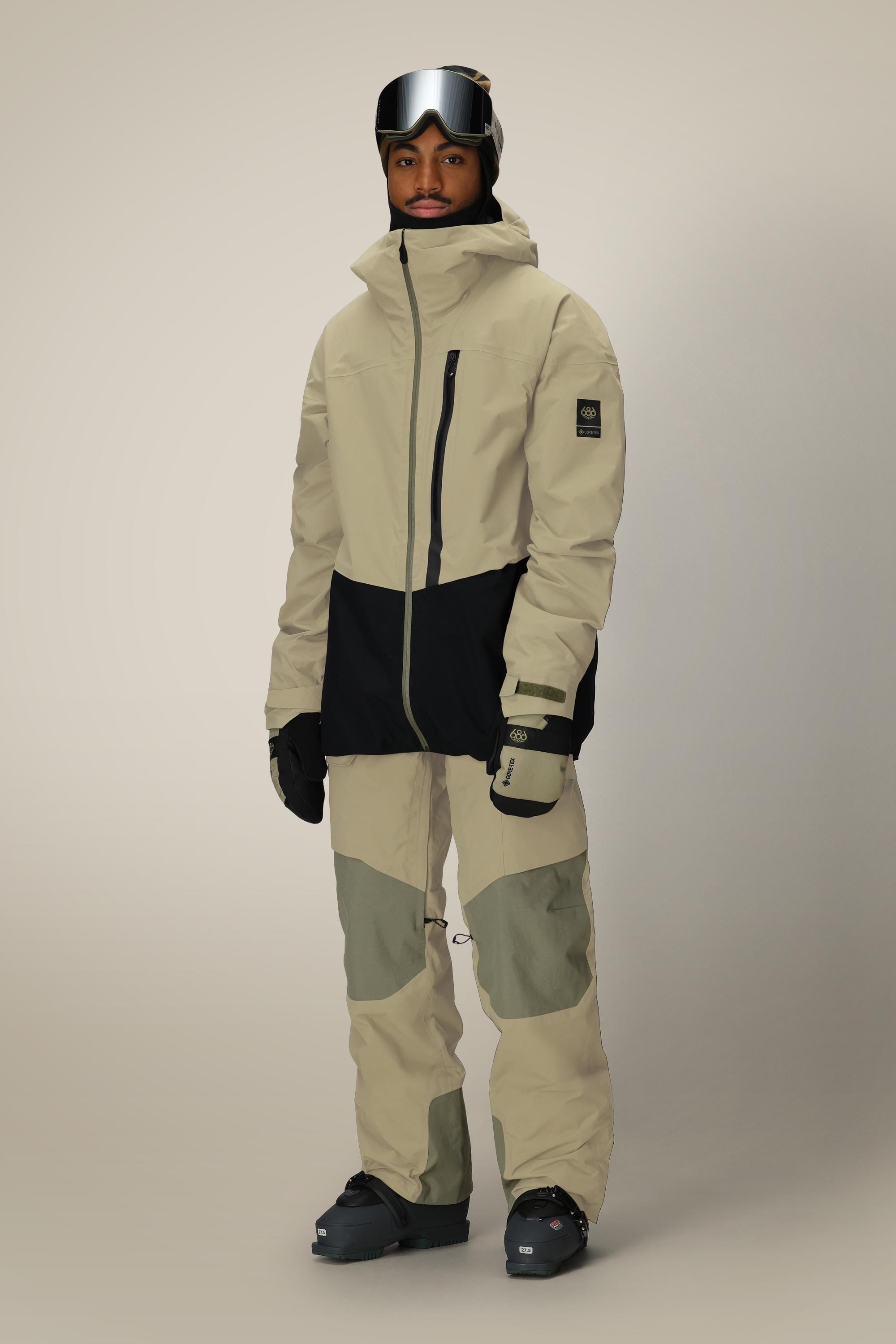A person stands facing forward against a plain background, wearing the 686 Men's GORE-TEX GT Thermagraph® Jacket in beige and black, paired with matching snow pants, gloves, black snow boots, and ski goggles.