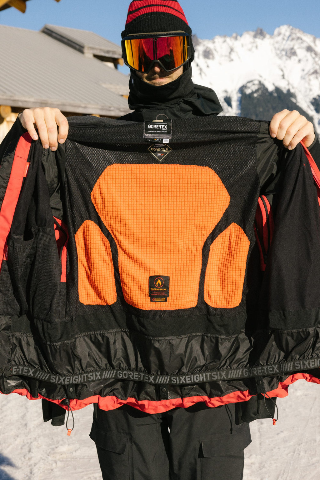 Wearing black winter gear and red goggles, a person displays the inner lining of the 686 Men's GORE-TEX GT Thermagraph® Jacket. Snowy mountains and a wooden building appear in the background.