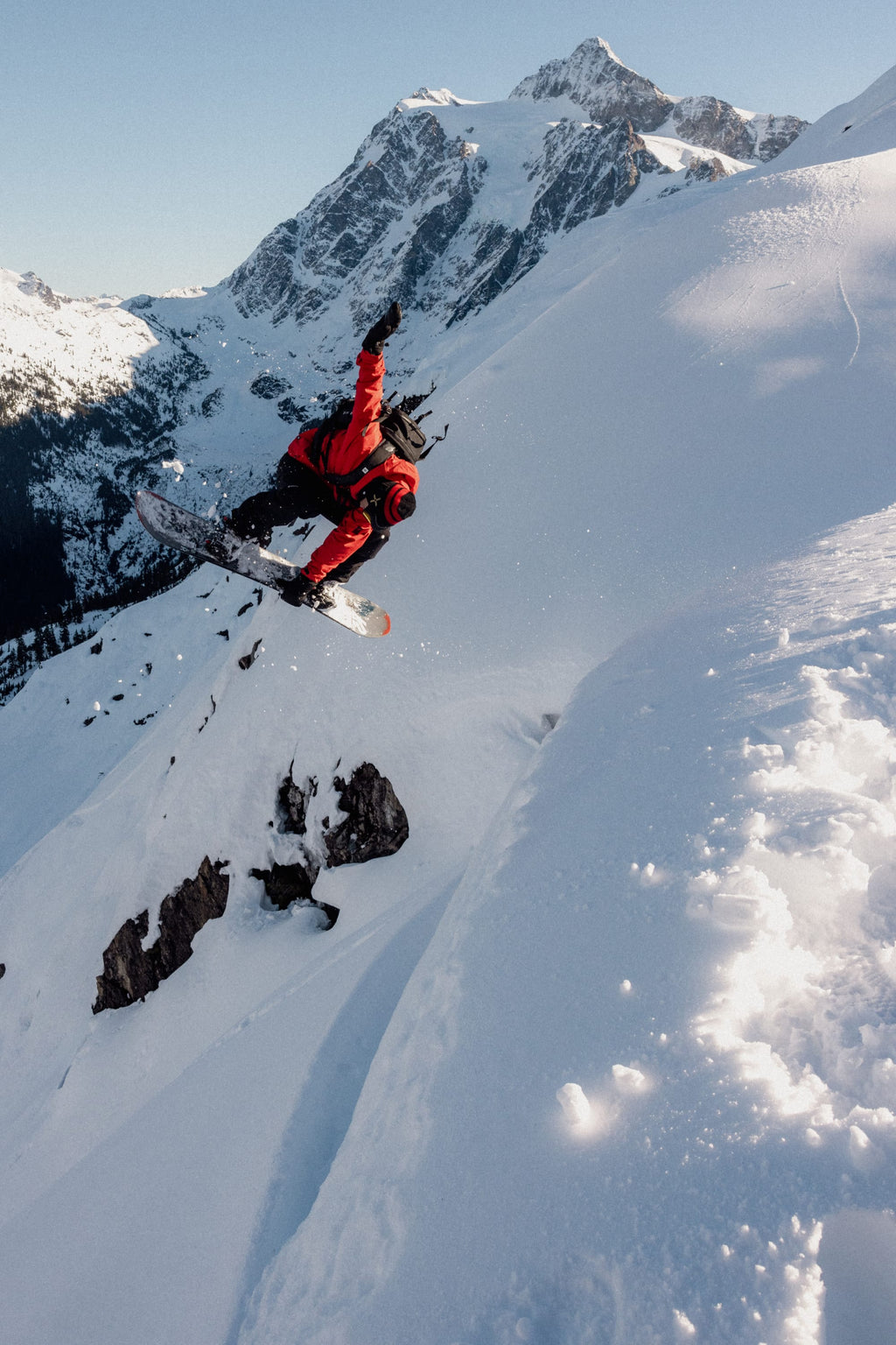 A snowboarder wearing the 686 Men's GORE-TEX GT Thermagraph® Jacket jumps off a snowy mountain cliff, with snow-covered peaks and a clear sky in the background.