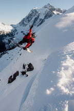 A snowboarder wearing the 686 Men's GORE-TEX GT Thermagraph® Jacket jumps off a snowy mountain cliff, with snow-covered peaks and a clear sky in the background.
