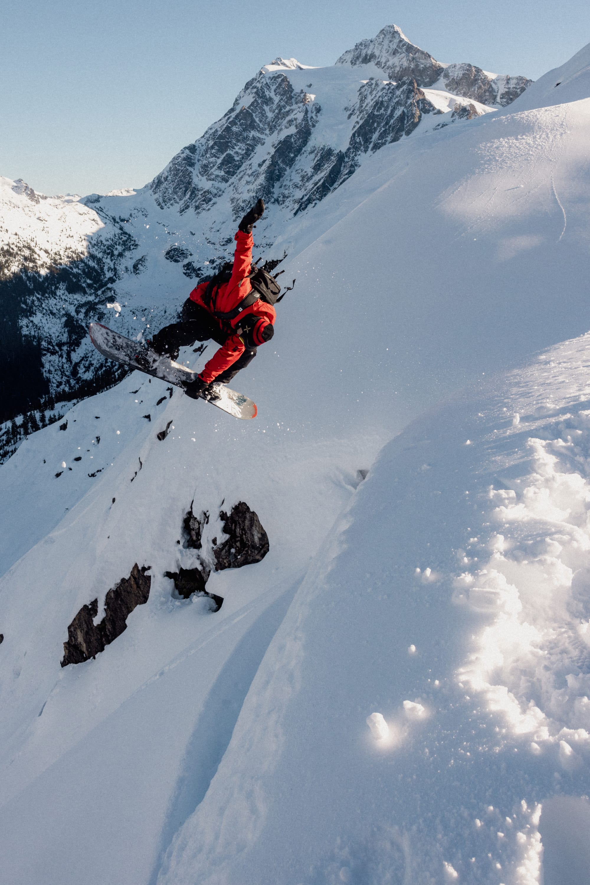 A snowboarder wearing the 686 Men's GORE-TEX GT Thermagraph® Jacket jumps off a snowy mountain cliff, with snow-covered peaks and a clear sky in the background.