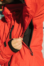 A person wearing the 686 Men's GORE-TEX GT Thermagraph® Jacket in red unzips an underarm vent, revealing a black inner layer. The scene is outdoors, possibly in snowy conditions.