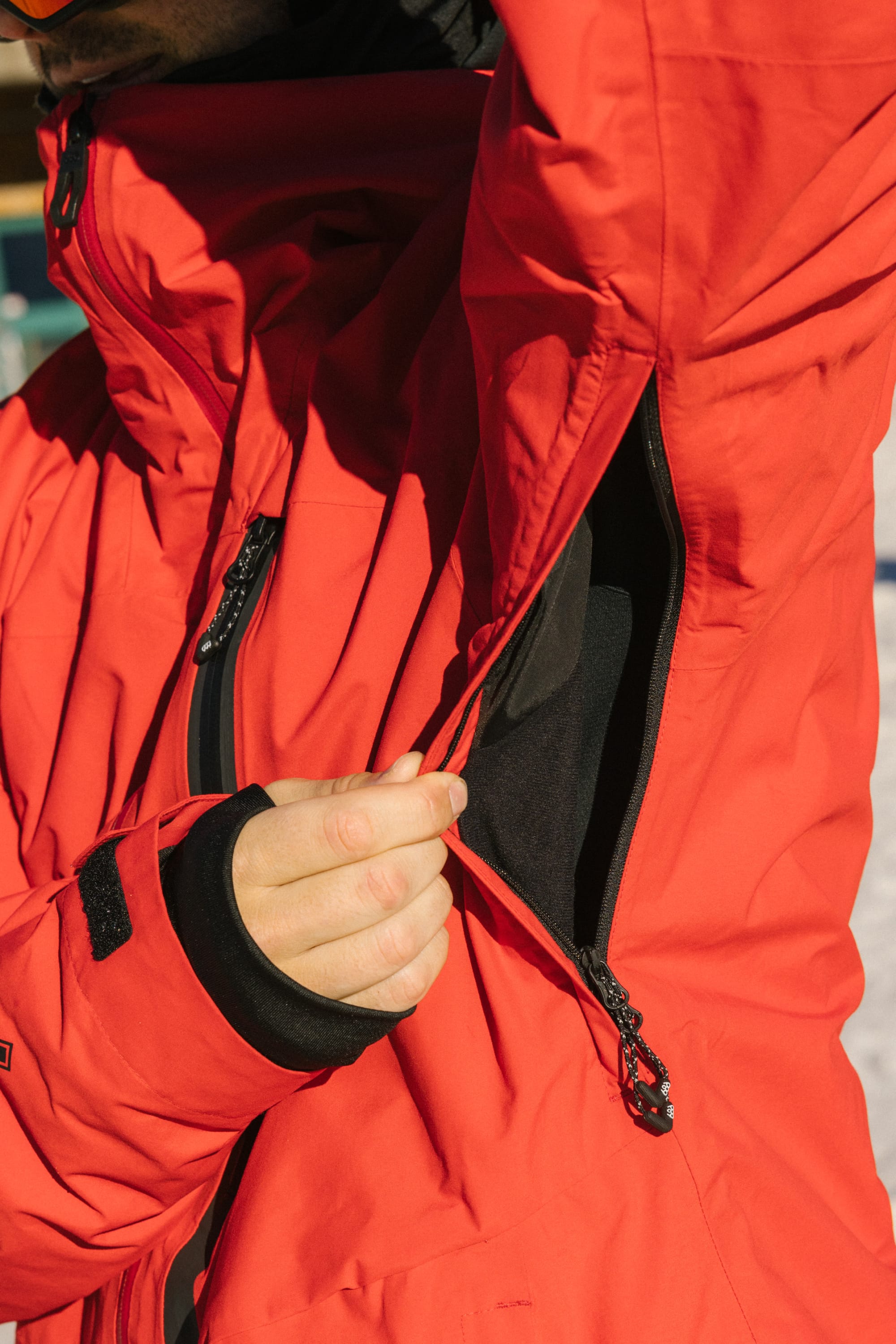 A person wearing the 686 Men's GORE-TEX GT Thermagraph® Jacket in red unzips an underarm vent, revealing a black inner layer. The scene is outdoors, possibly in snowy conditions.